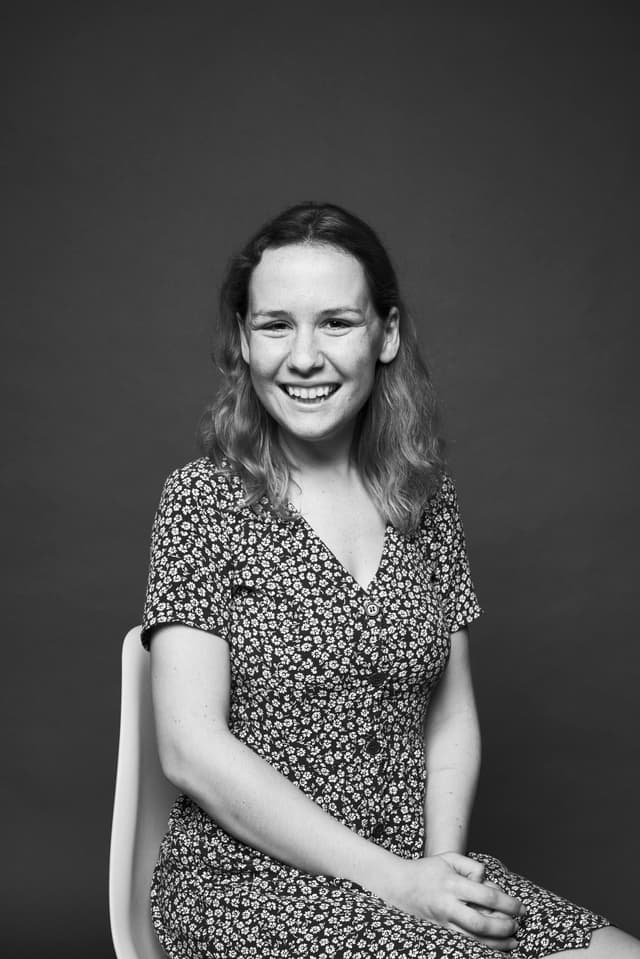 Black and white portrait of a smiling young woman with wavy hair, wearing a floral dress, seated on a modern chair against a plain background.