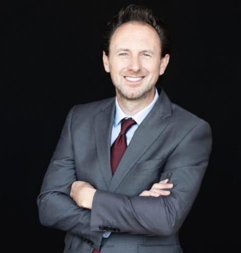 A smiling man in a gray suit with a red tie, arms crossed, standing against a black background.